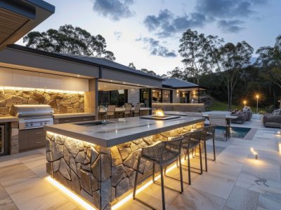 A modern outdoor kitchen with an island and barstools, set in the backyard of an Australian home at dusk. The kitchen features multiple grills, stone walls for cooking.